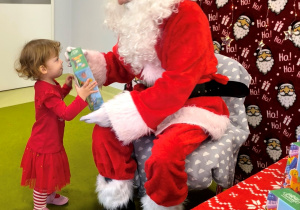 Św. Mikołaj siedzi na krześle. Rozalka podchodzi do Mikołaja i odbiera swój prezent.