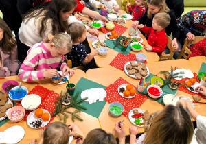 Dzieci i rodzice siedzą przy stolikach i wspólnie dekorują pierniczki.