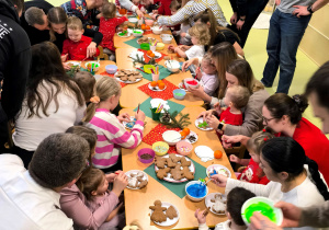 Dzieci i rodzice siedzą przy stolikach i wspólnie dekorują pierniczki i tworzą świąteczne dekoracje.