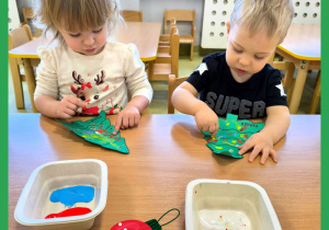 Dzieci siedzą przy stoliku i za pomocą paluszków i kolorowych farb dekorują choinki wycięte z papieru.