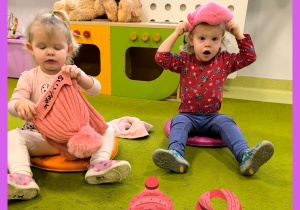 Dzieci siedzą na dywanie i samodzielnie próbują zakładać czapkę i szalik.