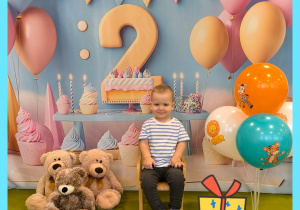 Adaś siedzi na krzesełku. za Adasiem powieszone jest urodzinowe tło. Obok stoją misie i kolorowe balony.