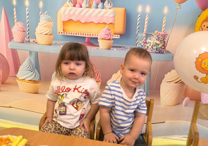 Adaś i Natalka siedzą przy stole. Za nimi powieszone jest urodzinowe tło.