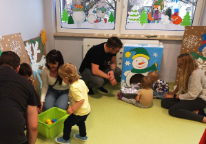 Dzieci z rodzicami bawią się piłeczkami i kulkami z papieru celując do bałwanka i choinki.