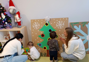 Dzieci z rodzicami bawią się wrzucając kolorowe piłki do otworów w dużej tekturowej choince.
