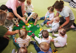 Dzieci z pomocą opiekunek przyklejają ozdoby do filcowej choinki.
