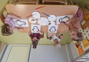 Dzieci siedzą przy stole i przyklejają watę na brzuszek pingwina.