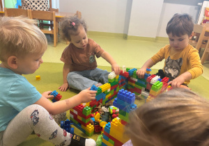 Dzieci siedzą na dywanie i układają zagrodę dla zwierząt z klocków.