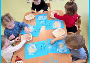 Dzieci siedzą przy stole i wykonują pracę plastyczną. Wyklejają za pomocą ryżu bałwana.