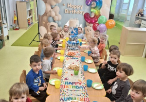 Dzieci siedzą przy stole na którym znajduje się urodzinowy poczęstunek.