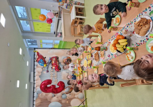 Dzieci siedzą przy stole na którym znajduje się urodzinowy poczęstunek.