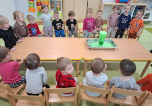 Dzieci obserwują przy stoliku doświadczenie z zielonym wybuchającym wulkanem.
