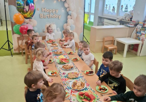 Dzieci siedzą przy stole na którym znajduje się urodzinowy poczęstunek.