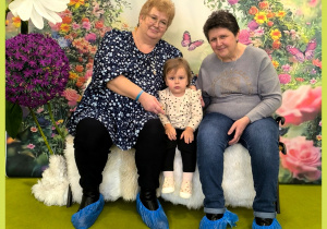 Julka i dwie babcie siedzą na ławeczce i pozują do zdjęcia. Za nimi powieszone jest tło z kwiatami.