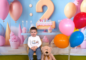 Dawidek siedzi na krześle i pozuje do urodzinowego zdjęcia z misiem i kolorowymi balonami.