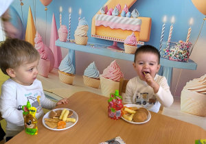 Dawidek i Franek siedzą przy stole i jedzą urodzinowy poczęstunek.