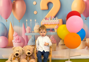 Staś siedzi na krześle i pozuje do urodzinowego zdjęcia z misiami i kolorowymi balonami.