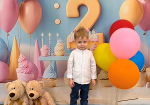 Staś stoi na dywanie i pozuje do urodzinowego zdjęcia z kolorowymi balonami.