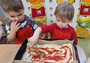 Antoni i Maksymilian smarują ciasto na pizzę sosem pomidorowym.