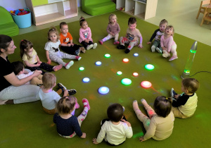 Dzieci z opiekunką siedzą na zielonym dywanie w kole i oglądają kolorowe światła żółty, zielony, różowy, czerwony, niebieski.