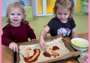 Pola i Antonina siedzą przy stole i przygotowują pizzę z okazji Międzynarodowego Dnia Pizzy.
