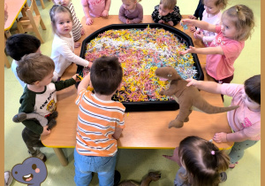 Dzieci stoją przy stole w kole i szukają zaginionych dinozaurów ukrytych w kolorowych ścinkach papieru.