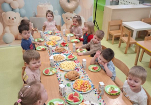 Dzieci siedzą przy stole na którym znajduje sie urodzinowy poczestunek.