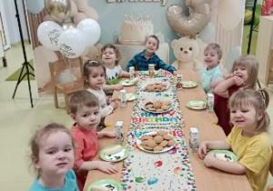 Dzieci siedzą przy stole na którym znajduje się urodzinowy poczęstunek