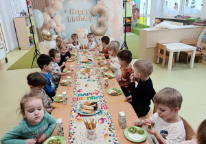 Dzieci siedzą przy stole na którym znajduje się urodzinowy poczęstunek.
