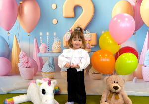 Marcelinka stoi i pozuje do urodzinowego zdjęcia na podłodze jest miś i jednorożec. Za Marcelinką powieszone jest urodzinowe tło z cyfrą dwa.