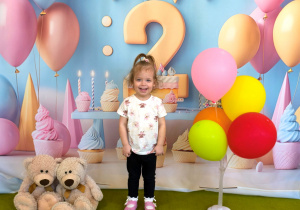 Rozalia stoi i pozuje do urodzinowego zdjęcia na podłodze są kolorowe balony i siedzą dwa misje. Za Rozalią jest powieszone urodzinowe tło z cyfrą dwa.