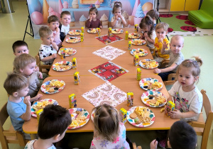 Wszystkie dzieci siedzą przy stole i jedzą urodzinowy poczęstunek.