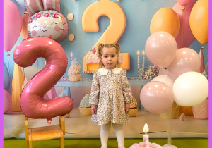 Zuzia stoi i pozuje do zdjęcia na tle urodzinowym. Obok stoją balony.