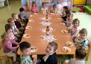 Dzieci siedzą przy stolikach i jedzą urodzinowy poczęstunek.