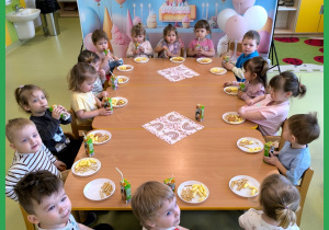 Dzieci siedzą przy stole i jedzą urodzinowy poczęstunek.