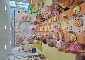 Dzieci siedzą przy stole na którym znajduje się urodzinowy poczęstunek.