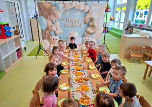 Dzieci siedzą przy stole na którym znajduje się urodzinowy poczęstunek.