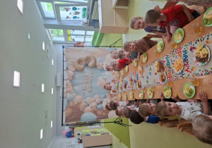Dzieci siedzą przy stole na którym znajduje się urodzinowy poczęstunek.