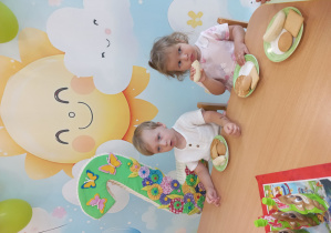 Ignacy i Amelia siedzą przy stole i jedzą urodzinowy poczęstunek za nimi jest tło urodzinowe ze słonkiem i cyfra dwa.