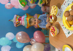 Jakub siedzi przy stole i pozuje do zdjęcia z balonami.