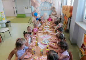 Dzieci siedzą przy urodzinowym stole i częstują się smakołykami.