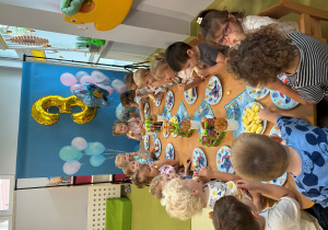 Dzieci siedzą przy stole i jedzą urodzinowy poczęstunek Marcelinki.