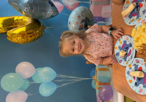 Marcelinka siedzi przy stole i pozuje do zdjęcia z balonami.