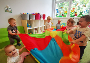 Dzieci z opiekunkami bawią się chustą animacyjną wykonując ruchy góra-dół oraz chowając się pod nią.