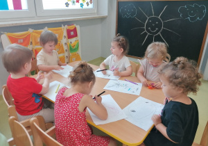 Dzieci siedzą przy stole i rysują kolorowymi kredkami.