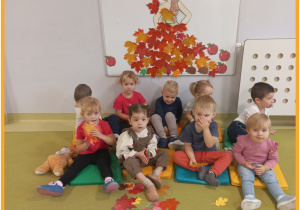 Dzieci siedzą na zielonym dywanie i pozują do zdjęcia w tle jest postać Pani Jesieni. ć