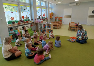Pani z biblioteki wraz z dziećmi siedzi na zielonym dywanie i czyta im książeczkę.