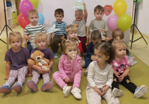 Dzieci siedzą na zielonym dywanie i pozują do grupowego zdjęcia w tle jest napis Dzień Chłopaka.