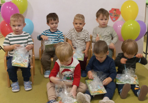 Chłopcy siedzą na zielonym dywanie i oglądają prezenty z okazji Dnia Chłopaka.