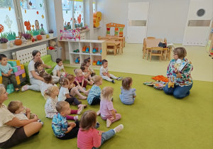Pani z biblioteki wraz z dziećmi siedzi na zielonym dywanie i czyta im książeczki.